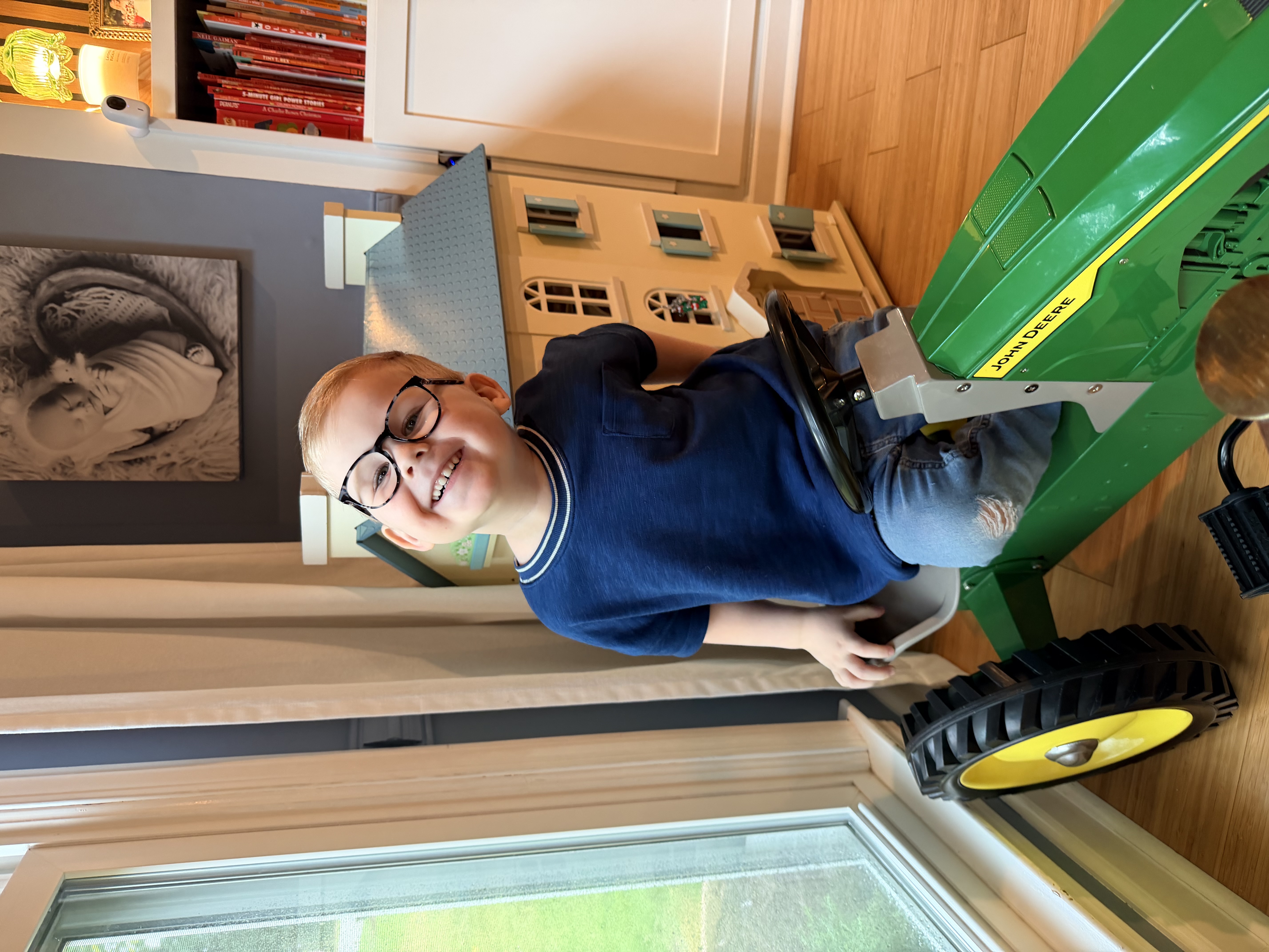 Felix grinning from the seat of his toy John Deere tractor at home